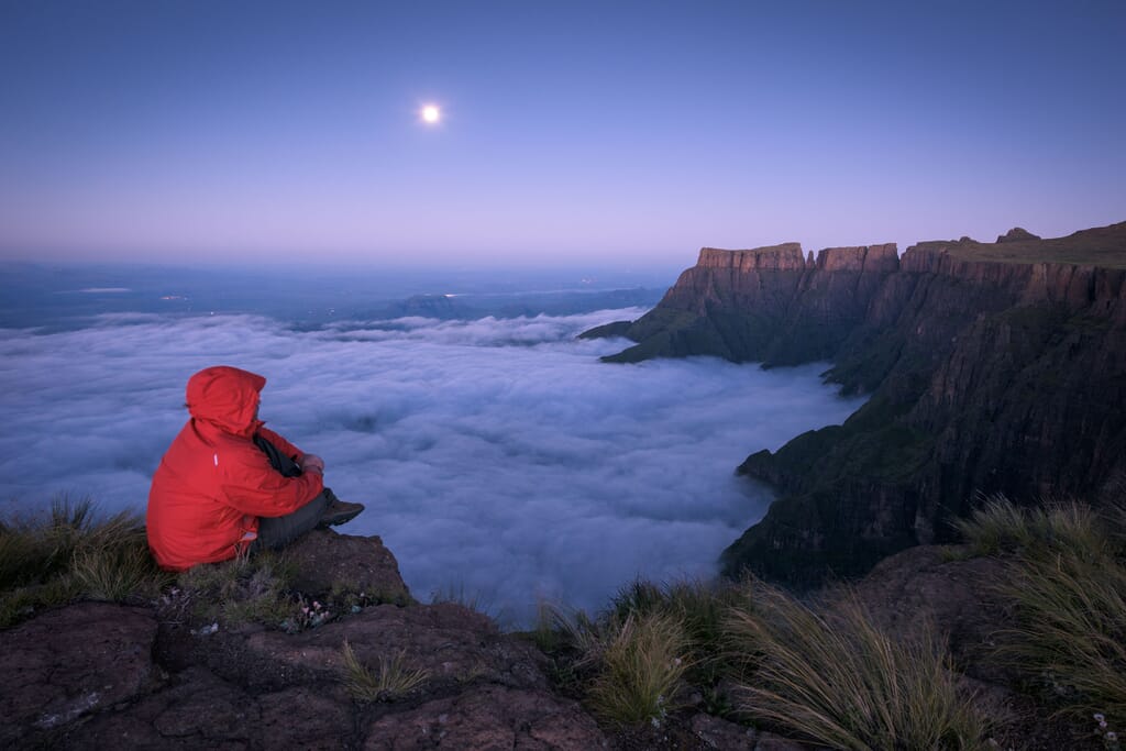 A horizontal landscape photograph of a lone man in a red jacket sitting on the edge of a cliff looking over the Drakensberg Amphitheatre mountains watching a full moon rise in South Africa.